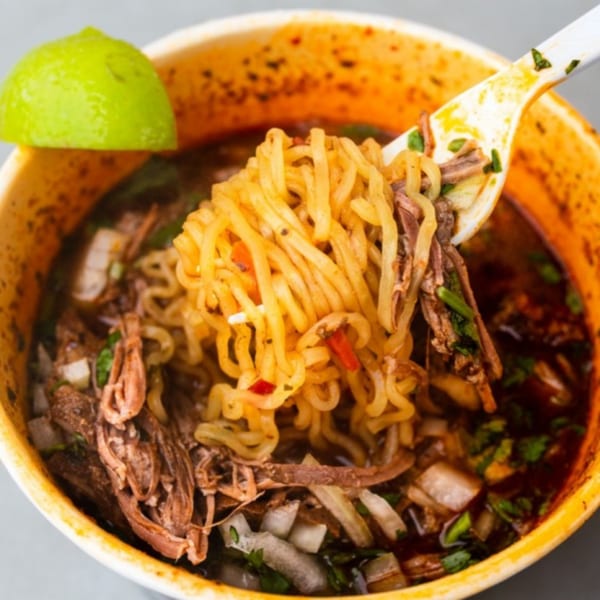 A close-up of birria ramen with shredded beef, noodles, and a lime wedge in a cup.