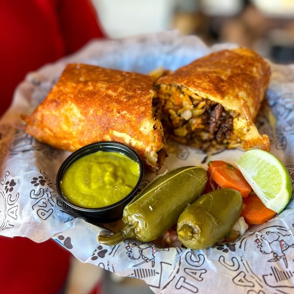 A Pacos Tacos burrito, cut in half, with a side of green sauce, pickled peppers, carrots, and lime.