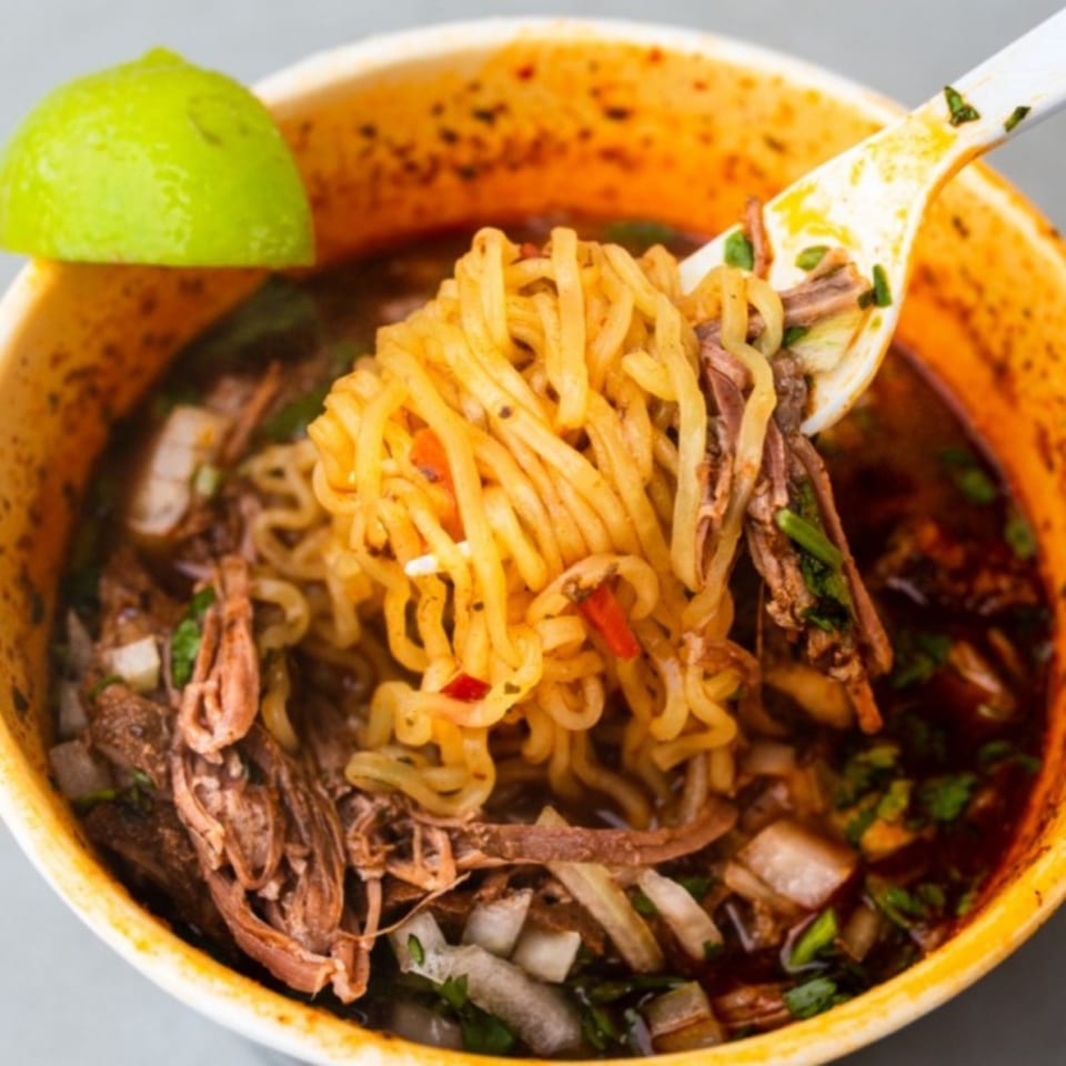 A close-up of birria ramen with shredded beef, noodles, and a lime wedge in a cup.