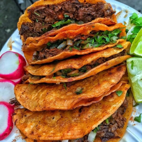 A plate of Pacos Tacos birria tacos with cilantro, onions, lime wedges, and radishes.